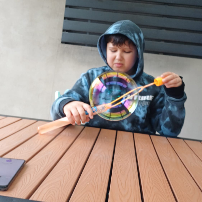 NDIS Child in a blue hoodie sitting at a wooden table, concentrating while playing with a colorful spinning bubble wand toy.